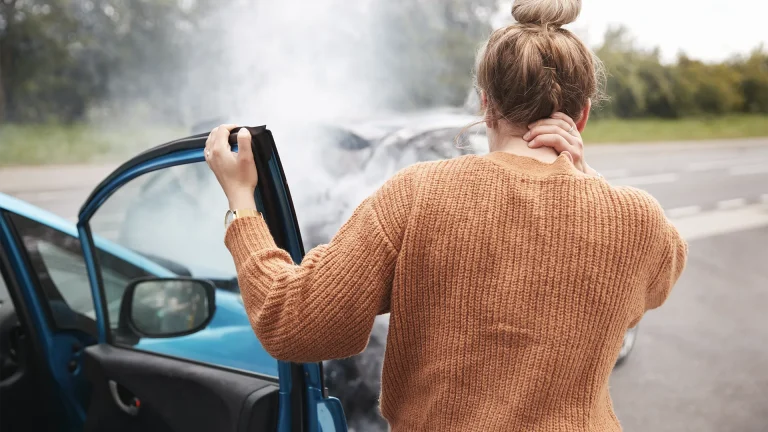 A woman is rubbing her neck after rear-ending a car in front of her. The collision damaged both vehicles and smoke is spreading around the engine.