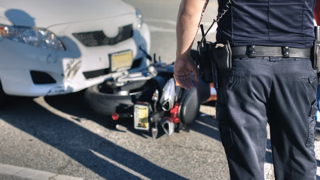Policeman overlooking a wreck involving a white car and a motorcycle down to the ground.