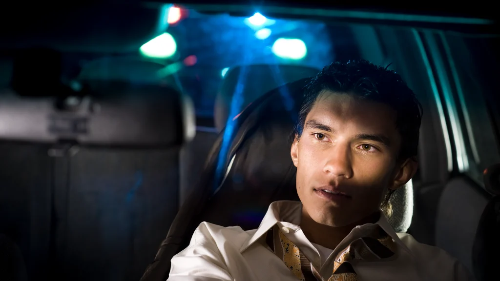 man sitting in car looking into rear view mirror with police lights flashing