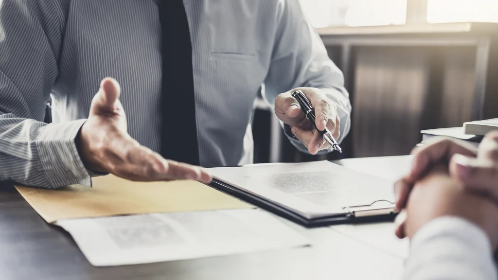 man in shirt and ties gesturing toward client with pen in hand at desk